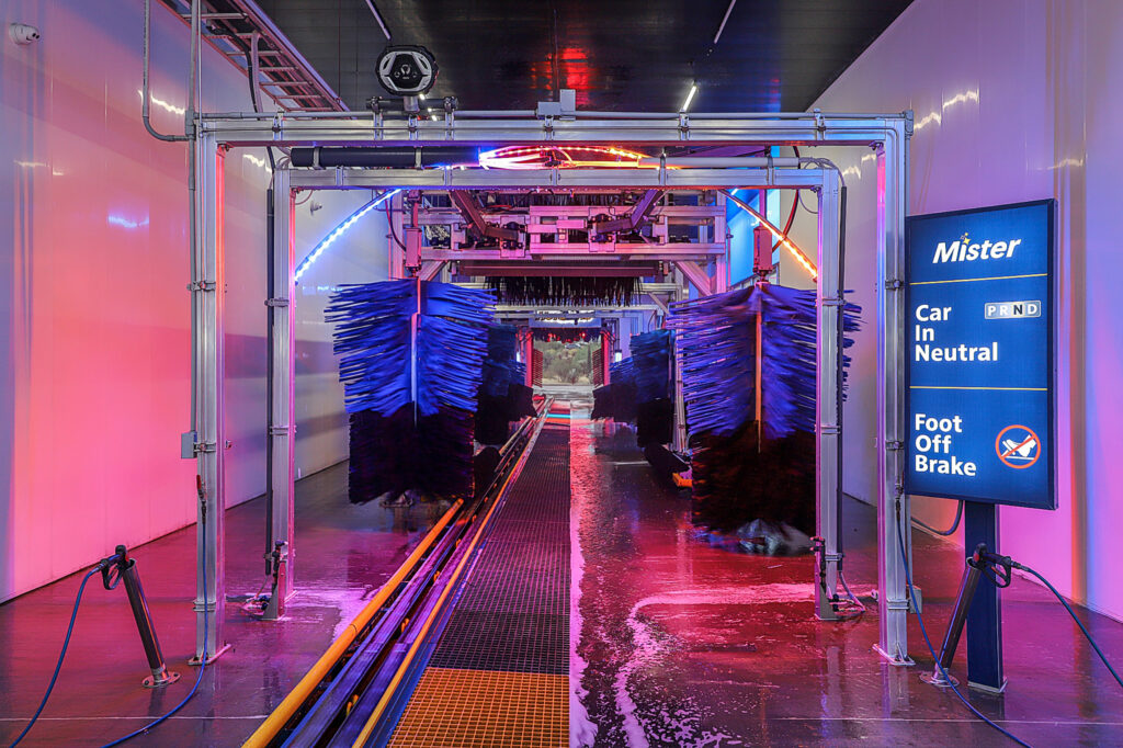 Interior of a commercial car wash tunnel with pink and blue lights and large car brushes.